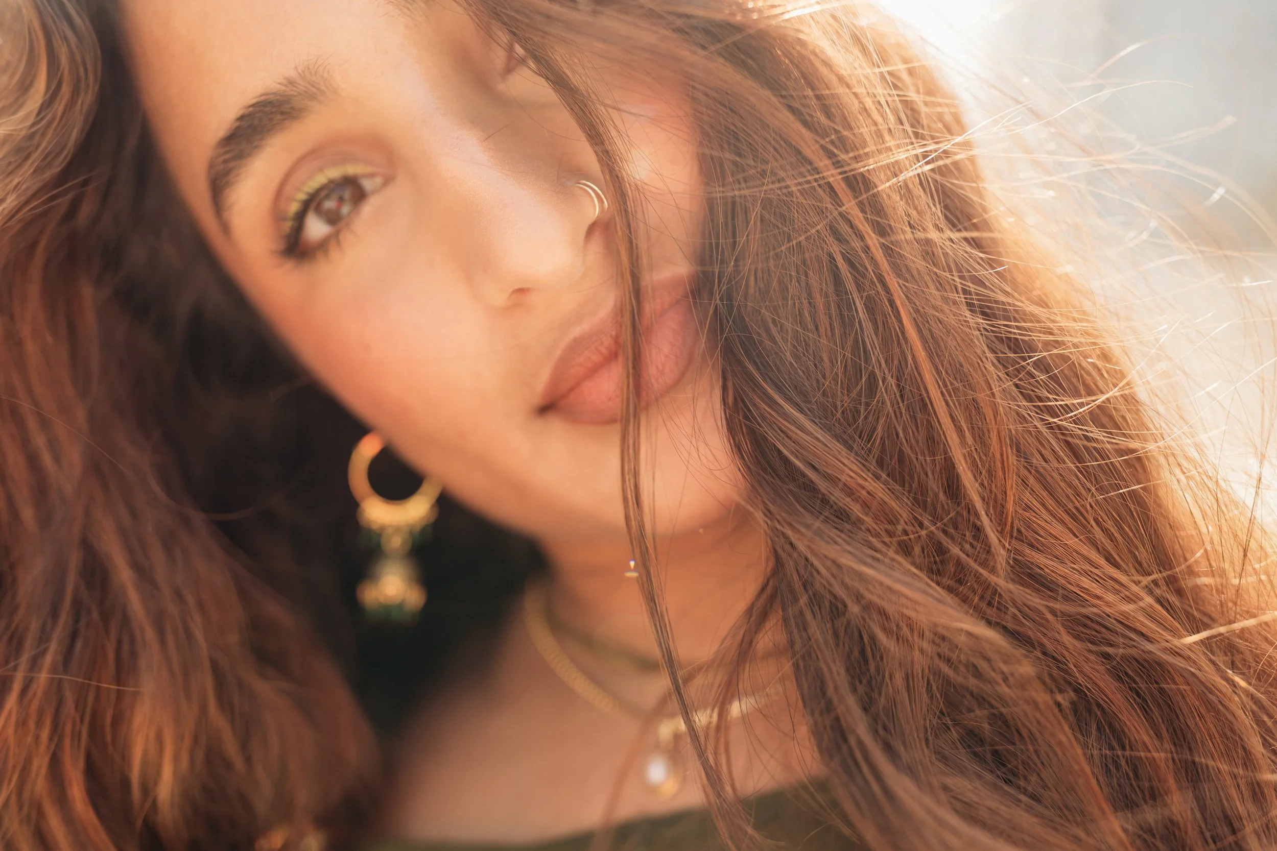 Close-up of a young woman with long, wavy hair, wearing gold hoop earrings and layered necklaces, smiling softly in warm sunlight.