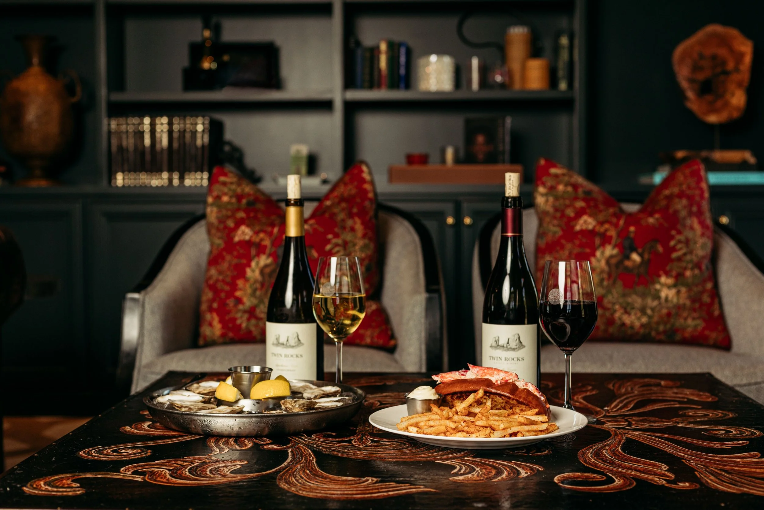 A table set with seafood, a lobster roll with fries, and two wine bottles with glasses of white wine and red wine, in a cozy room with decorative pillows and a dark bookshelf in the background.