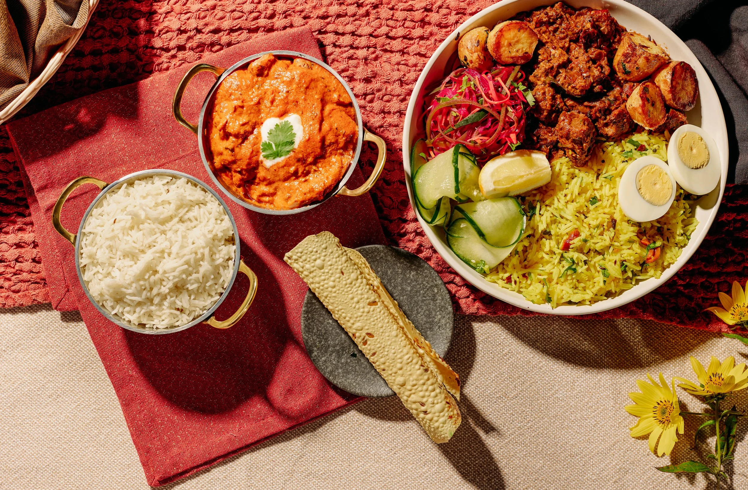 An Indian meal spread featuring a large bowl of yellow rice, a bowl of curry, a bowl of white rice, a slice of naan bread on a black plate, and small servings of onion salad, lemon, and cucumber slices.