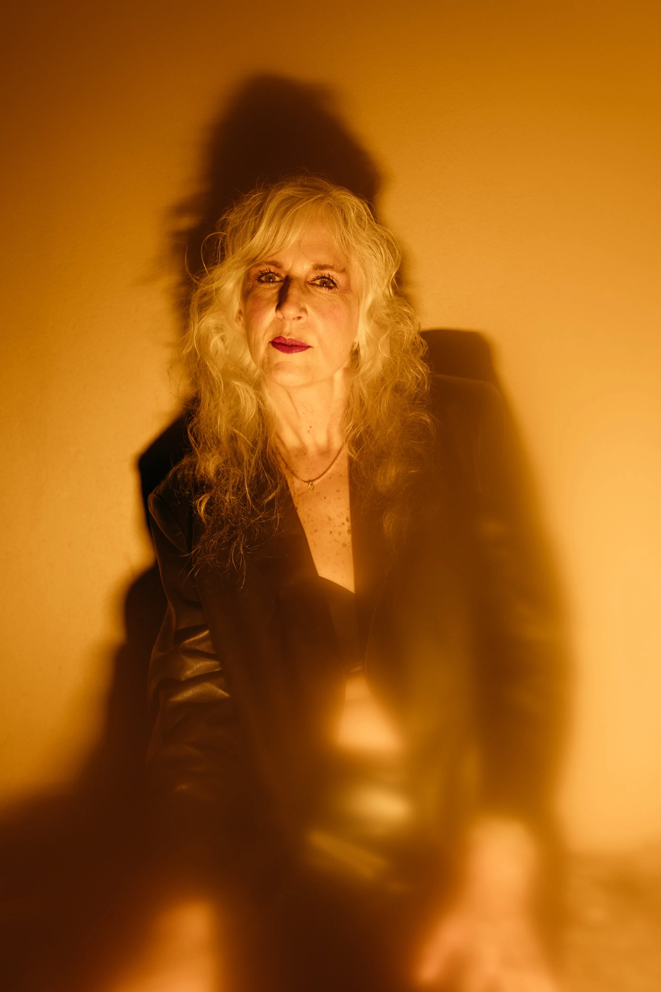 A woman with long, curly blonde hair and fair skin, wearing a black outfit, sitting against a beige wall. The lighting creates a prominent shadow of her head and upper body on the wall behind her.