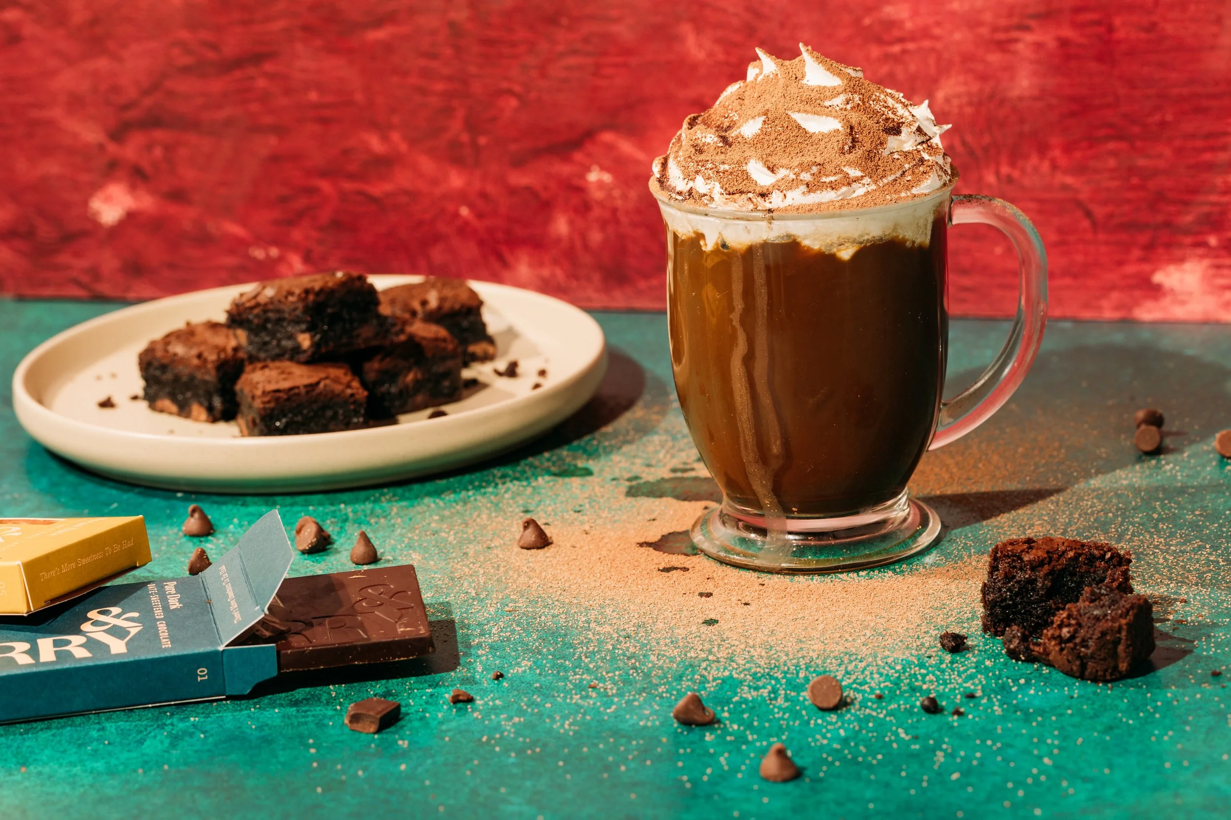 A glass mug filled with a hot beverage topped with whipped cream and cocoa powder, surrounded by chocolate brownies, broken chocolate bars, and scattered chocolate chips on a green surface with a red background.