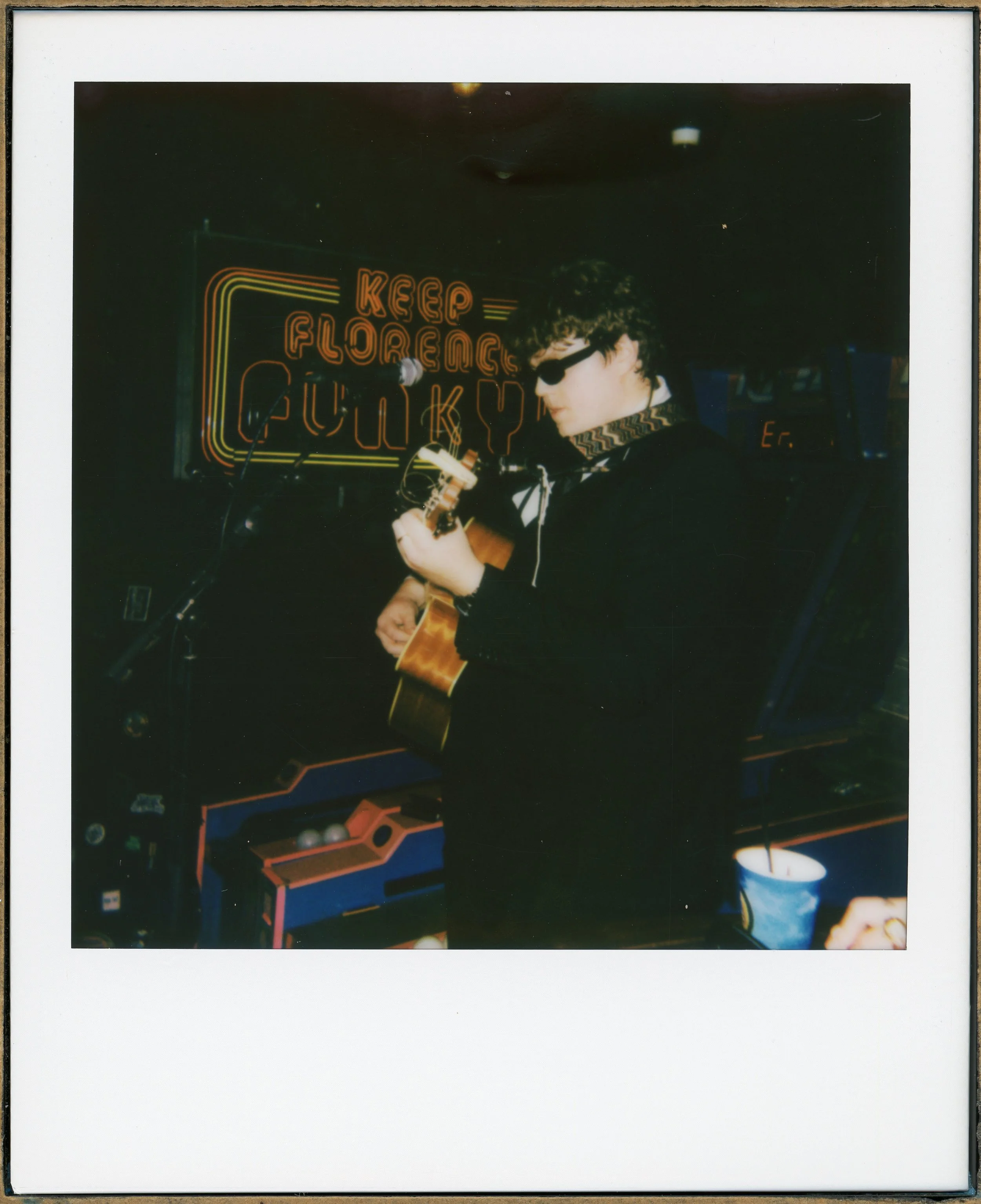 A person wearing sunglasses and a dark outfit playing an acoustic guitar. Behind them is a neon sign that reads 'Keep Flipping Funky' and a vintage arcade game machine.