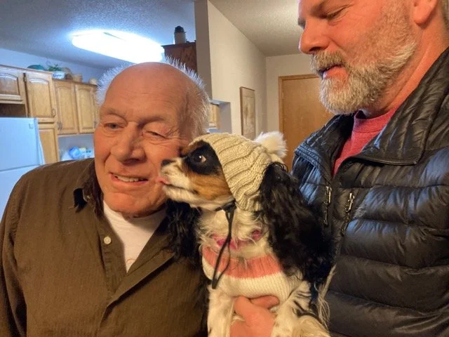 An elderly man and a middle-aged man are holding a small dog between them. The elderly man is smiling and winking, while the dog is wearing a knit hat and is licking the elderly man's face. The setting is inside a kitchen or living area.