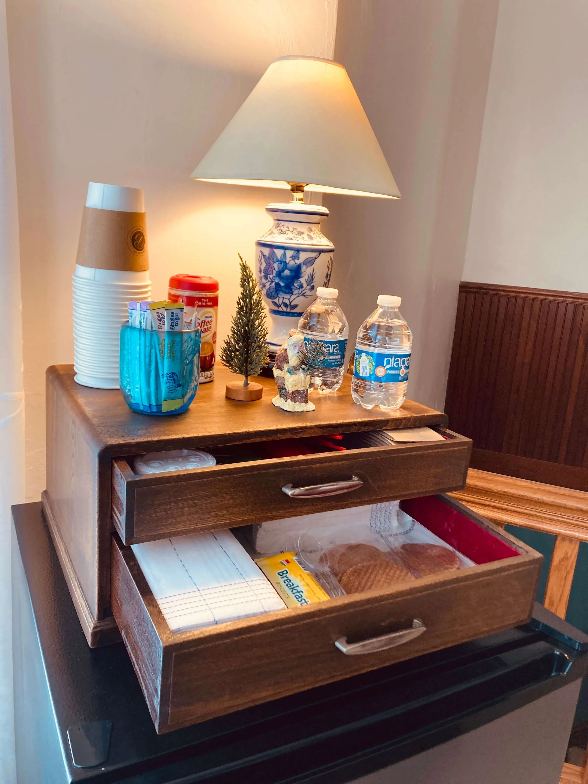 Wooden breakfast tray with two open drawers containing plates and bread, placed on top of a black surface, with a lamp, water bottles, cups, and small decorative items on it.