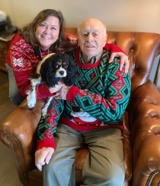A woman and an elderly man are sitting on a brown leather chair, smiling at the camera. The woman has shoulder-length brown hair and is wearing a red holiday sweater, while the man has light hair and is wearing a Christmas sweater with green, red, an