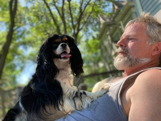 Man lying outside with a Cavalier King Charles Spaniel sitting on his chest, outdoors on a sunny day with green trees in the background.