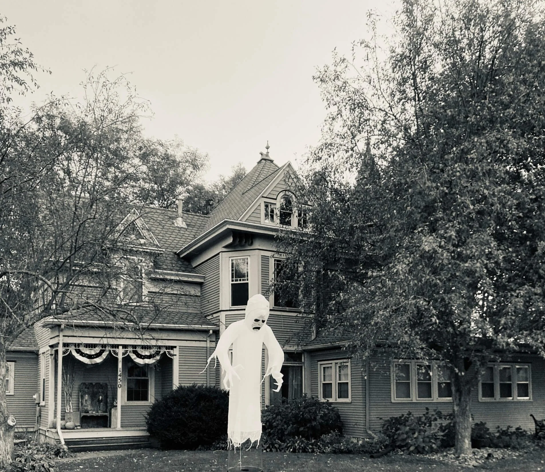 A Halloween decoration of a ghost with a scary face and tattered sheet stands in front of a large vintage house with trees and bushes around.