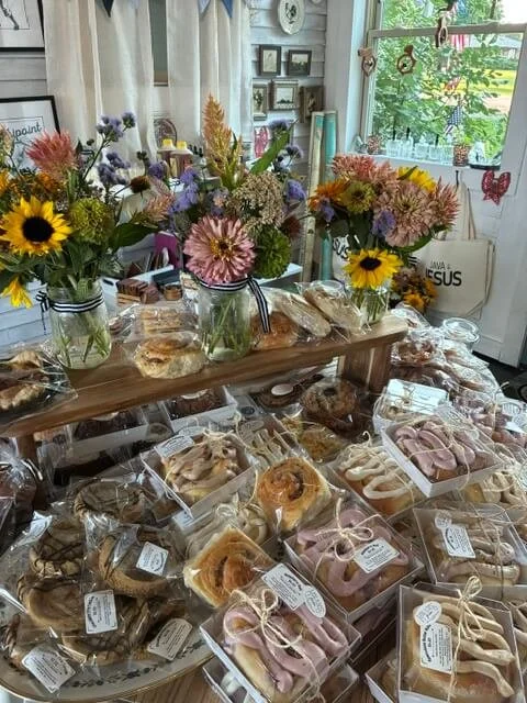 A table with bakery items like cookies and cakes, decorated with colorful flower arrangements and a window in the background.