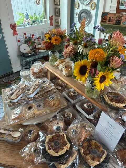 A table with a variety of baked goods wrapped in plastic, including cookies, muffins, and fruit tarts. There is a large bouquet of colorful flowers in a vase, with sunflowers, daisies, and other flowers. In the background, a white dresser decorated w