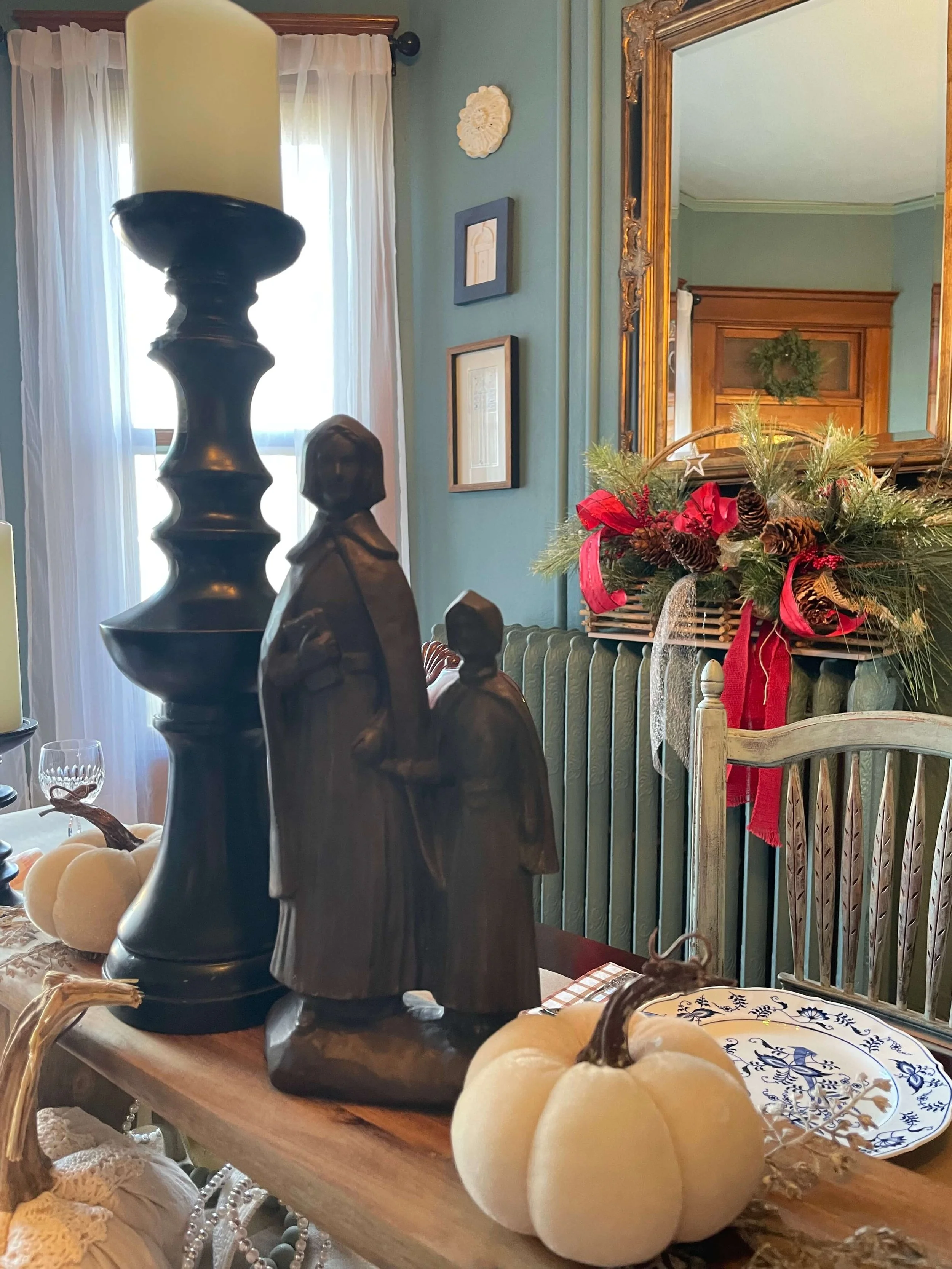 Decorative autumn-themed display featuring small white pumpkins, a dark wooden angel figurine, and a bronze sculpture of a woman and a child in a dining room with framed artwork, a large mirror, and a holiday arrangement.