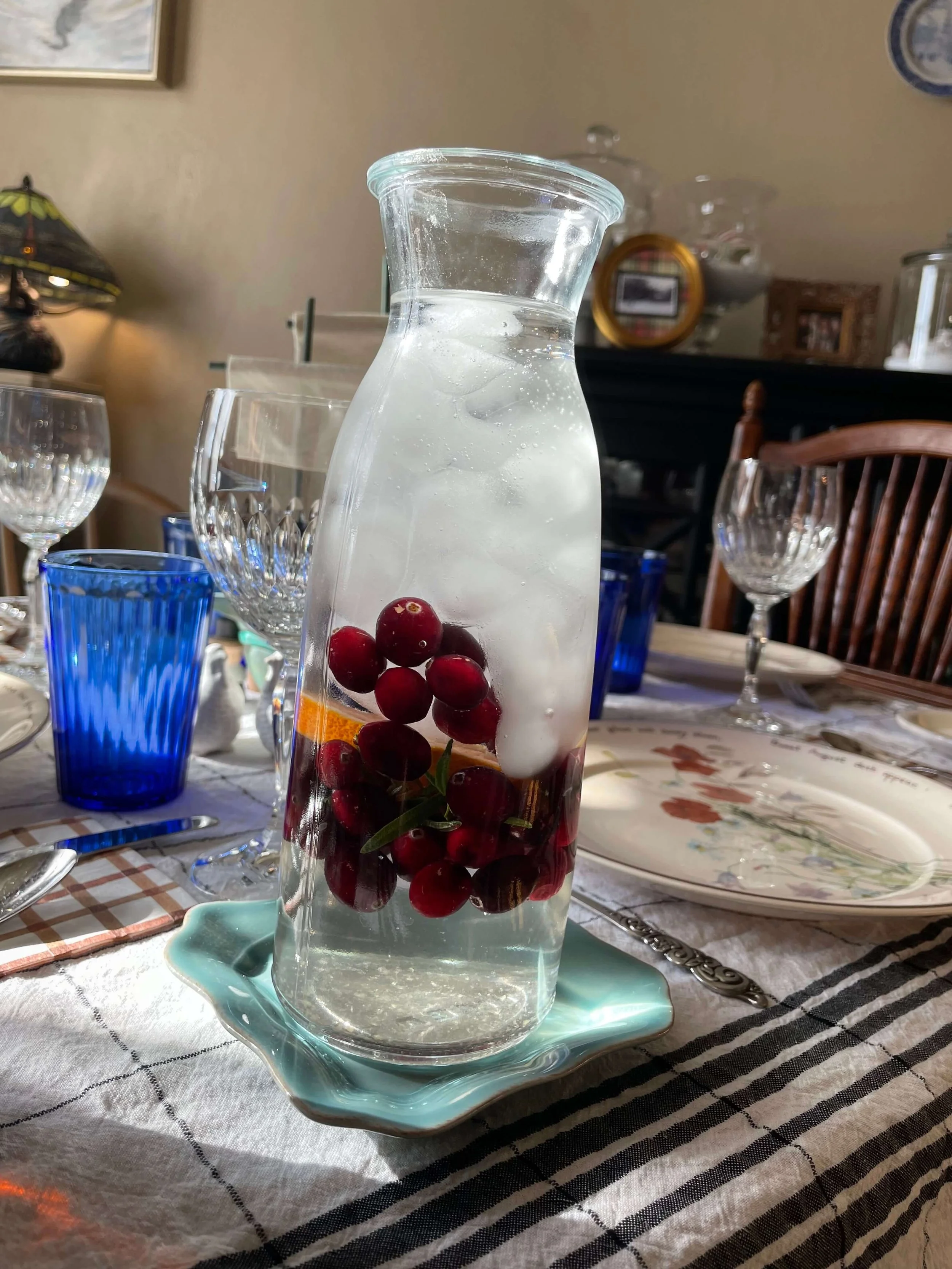 A glass pitcher filled with water, ice cubes, and red berries on a dining table surrounded by glasses, plates, and utensils.