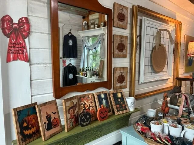 Wall decorated with framed art and Halloween-themed paintings, including pumpkins and a black cat. A red bow decoration with text is on the left. A mirror and various small jars and items are on a shelf below the framed art. A circular tray with misc
