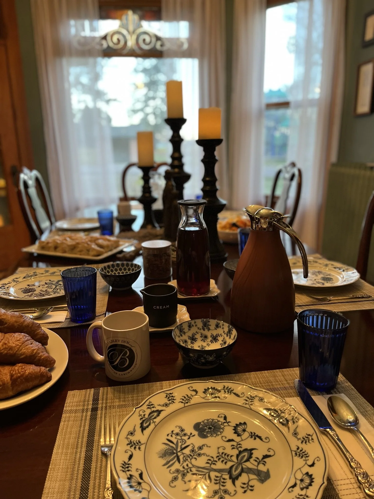 Dining table set with plates, cups, glasses, and food, with a chandelier above and windows in the background.