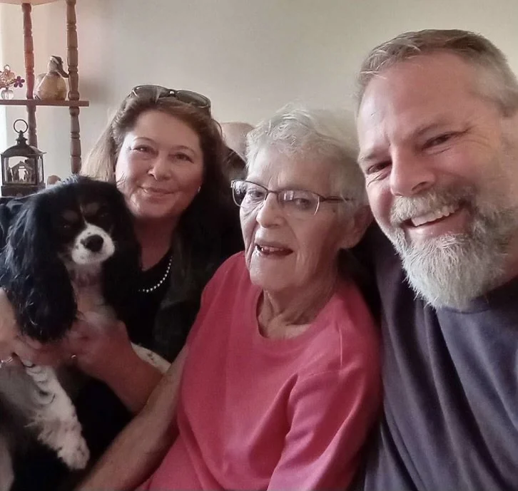 Four people and a dog posing together indoors, with a woman in a pink shirt at the center, an elderly woman with glasses, a woman with shoulder-length hair, and a man with a beard.