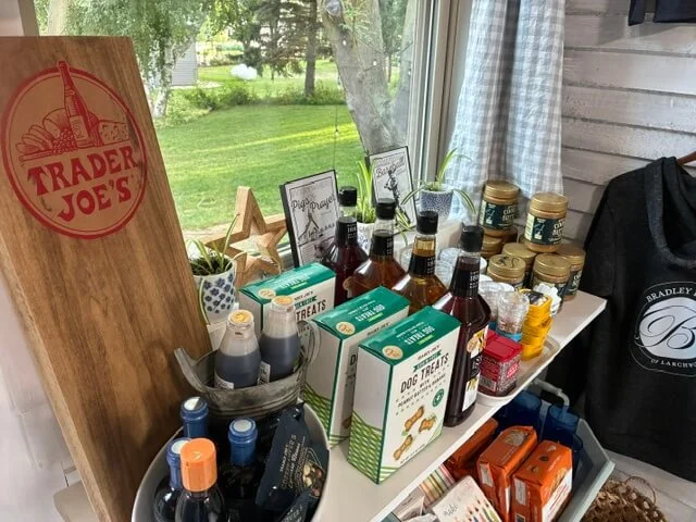 Display of Trader Joe's products including dog treats, bottled beverages, jars of condiments, and boxes on a table near a window.