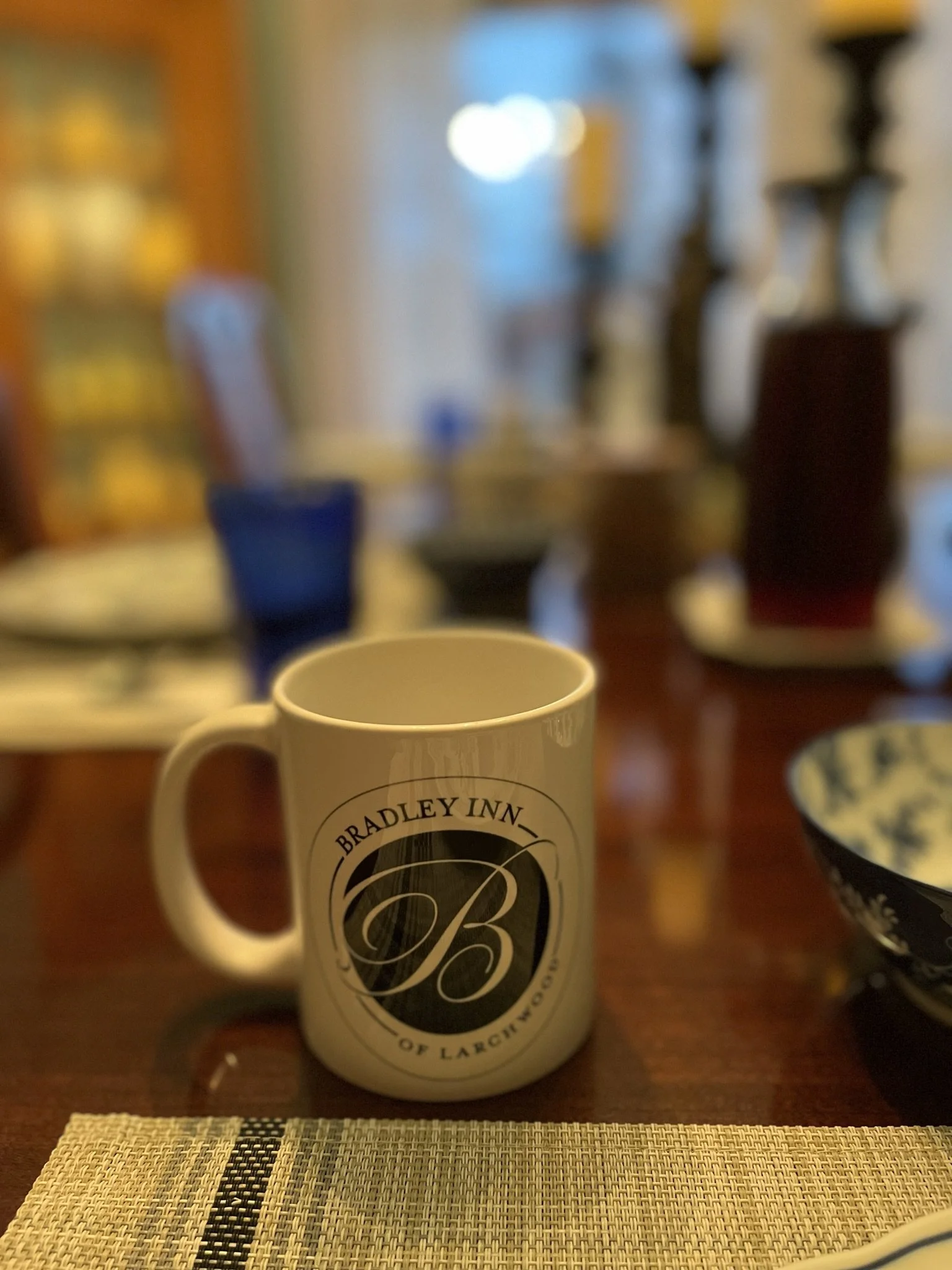 A white coffee mug with black and white logo reading 'Bradley Inn of La Crosse' on a wooden table, with a placemat in the foreground and a blurred background of a dining room with lights and furniture.