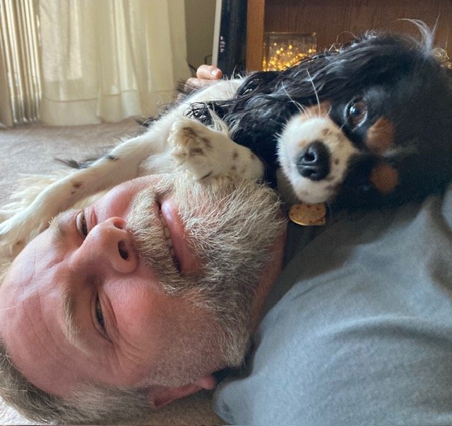 A man with gray hair and a beard lying on his back, smiling with closed eyes, snuggling with a small dog on his neck. The dog has black, white, and brown fur and is resting its head on the man's face, looking at the camera.