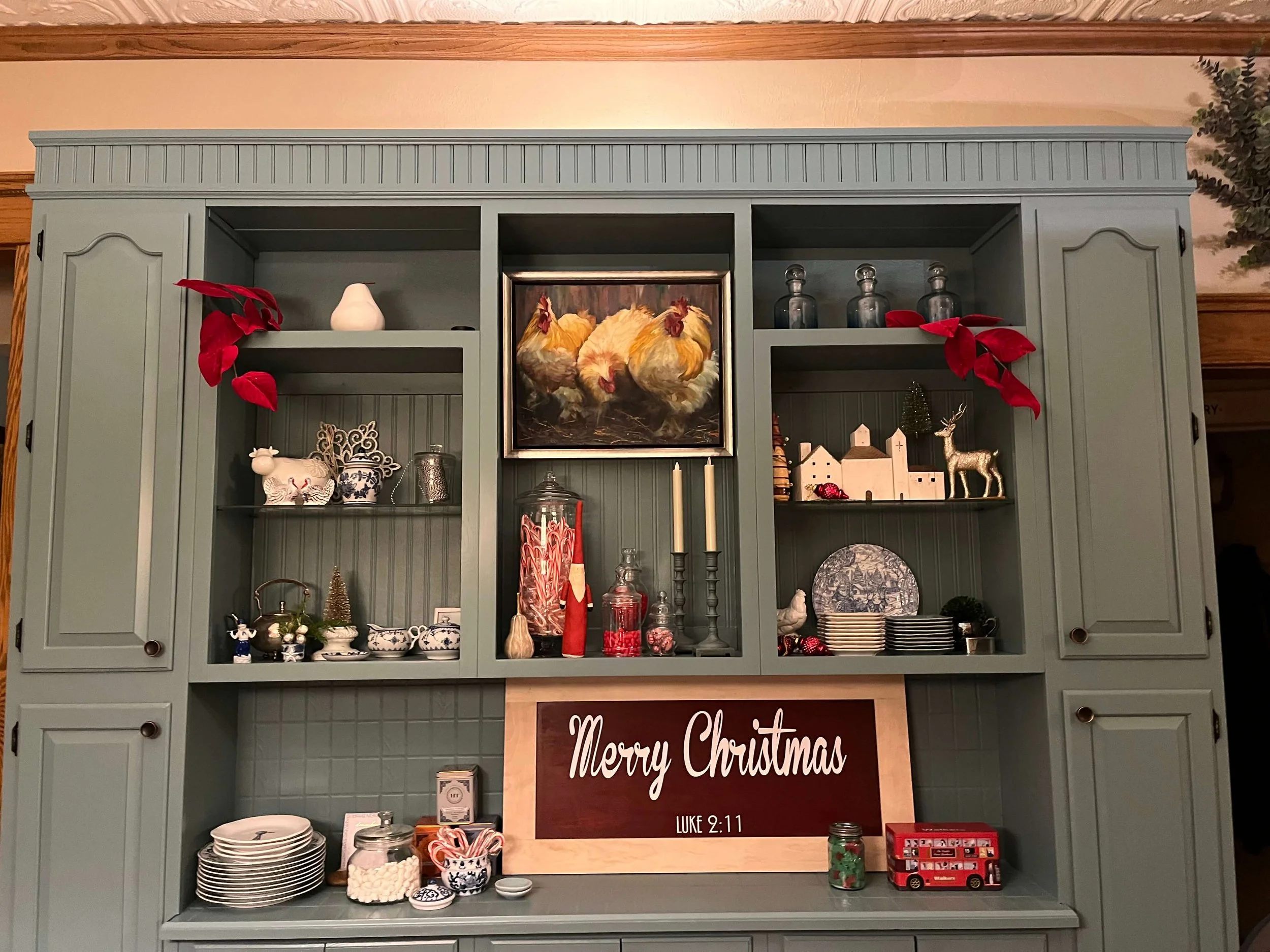 A green kitchen hutch decorated for Christmas with various holiday ornaments, including a sign that reads "Merry Christmas" with a Bible verse reference, and a painting of chickens above it.