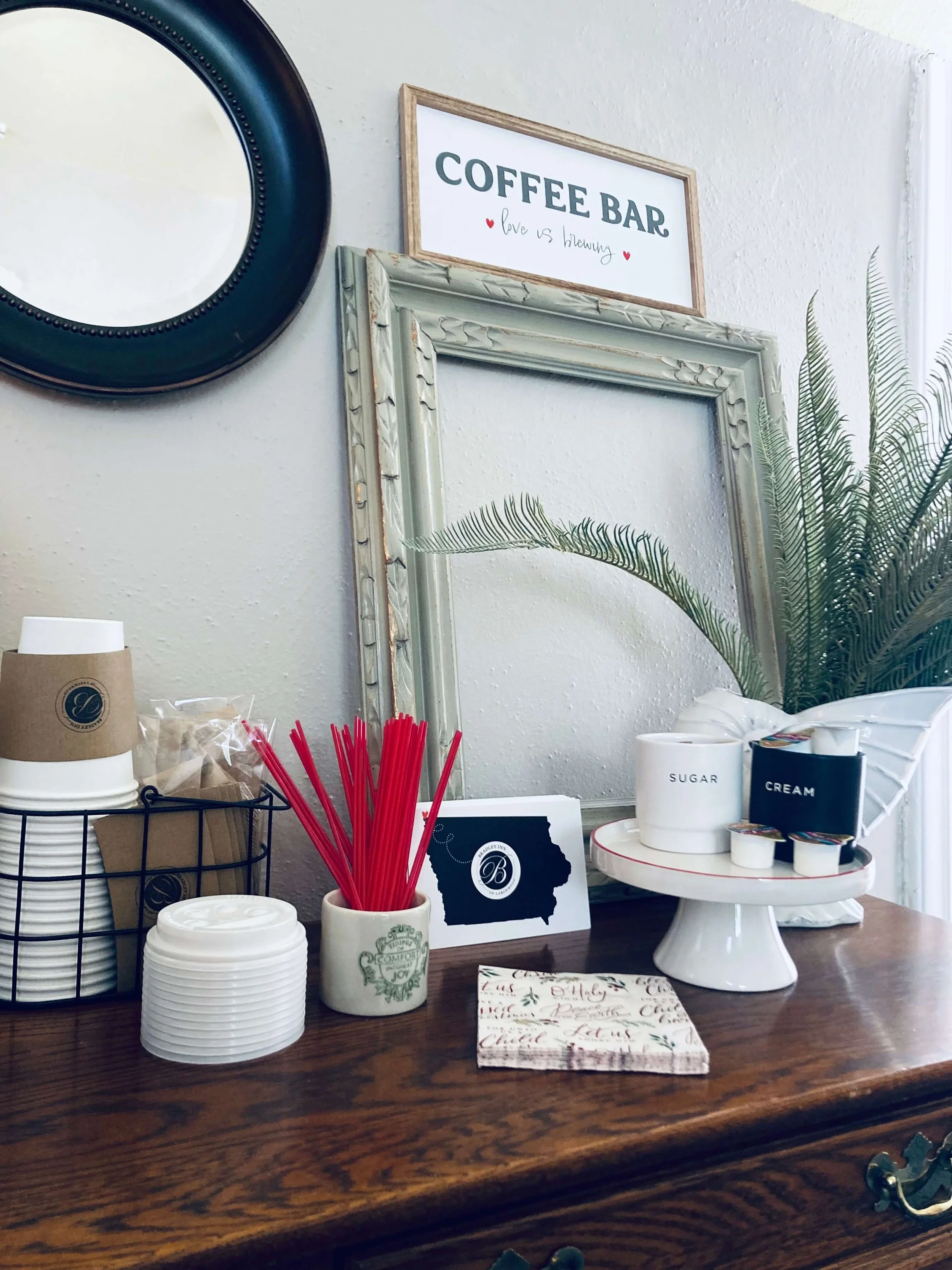 A coffee bar setup with disposable cups, red stir sticks, sugar and creamer containers, and a decorative napkin on a wooden table. There are framed mirrors and a sign reading 'COFFEE BAR' with the phrase 'love is brewing' on the wall behind.