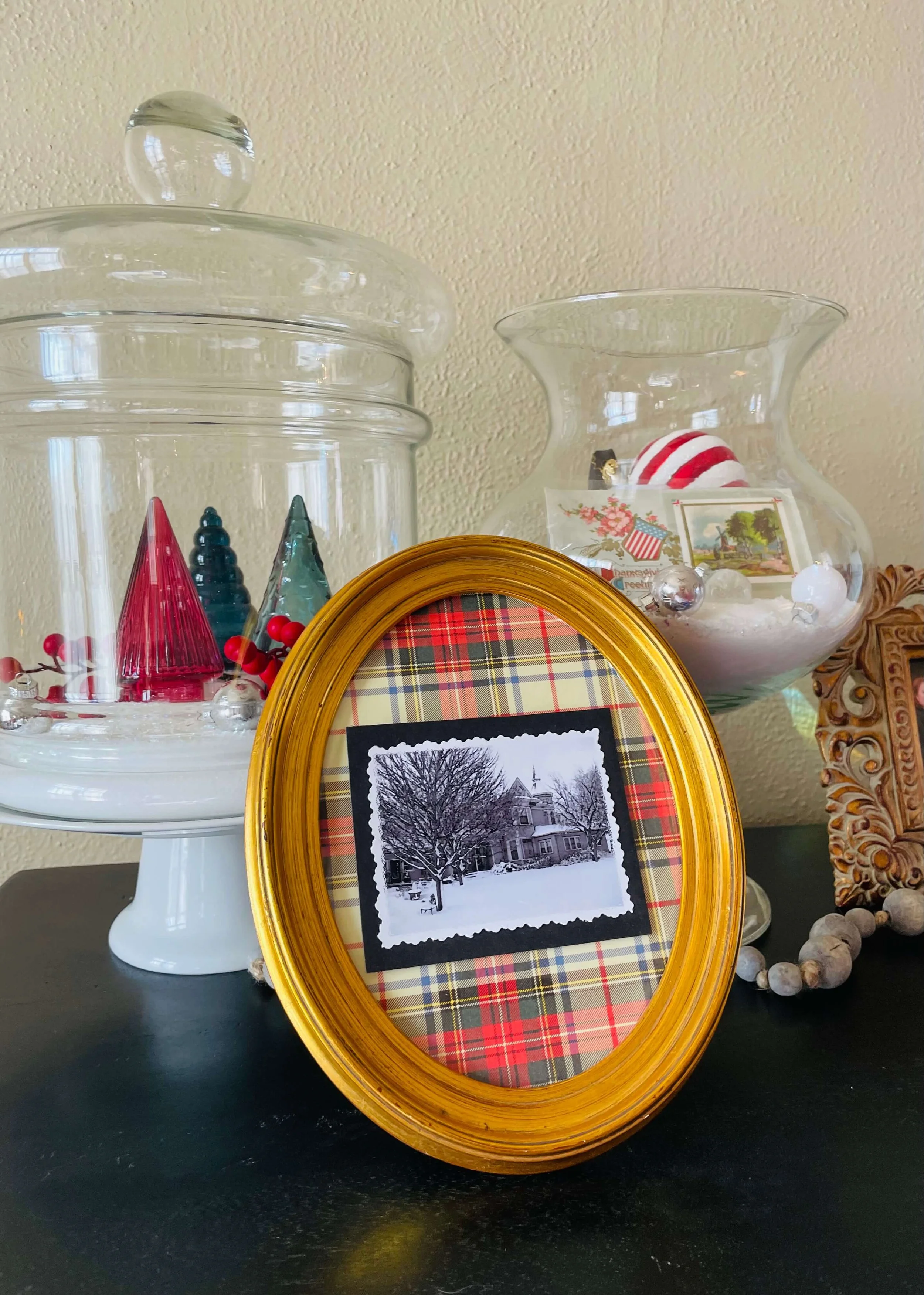 Decorative holiday items including glass jars with Christmas ornaments and figurines, a framed black and white photo of a snowy house, and a beaded garland, set on a dark surface against a light wall.