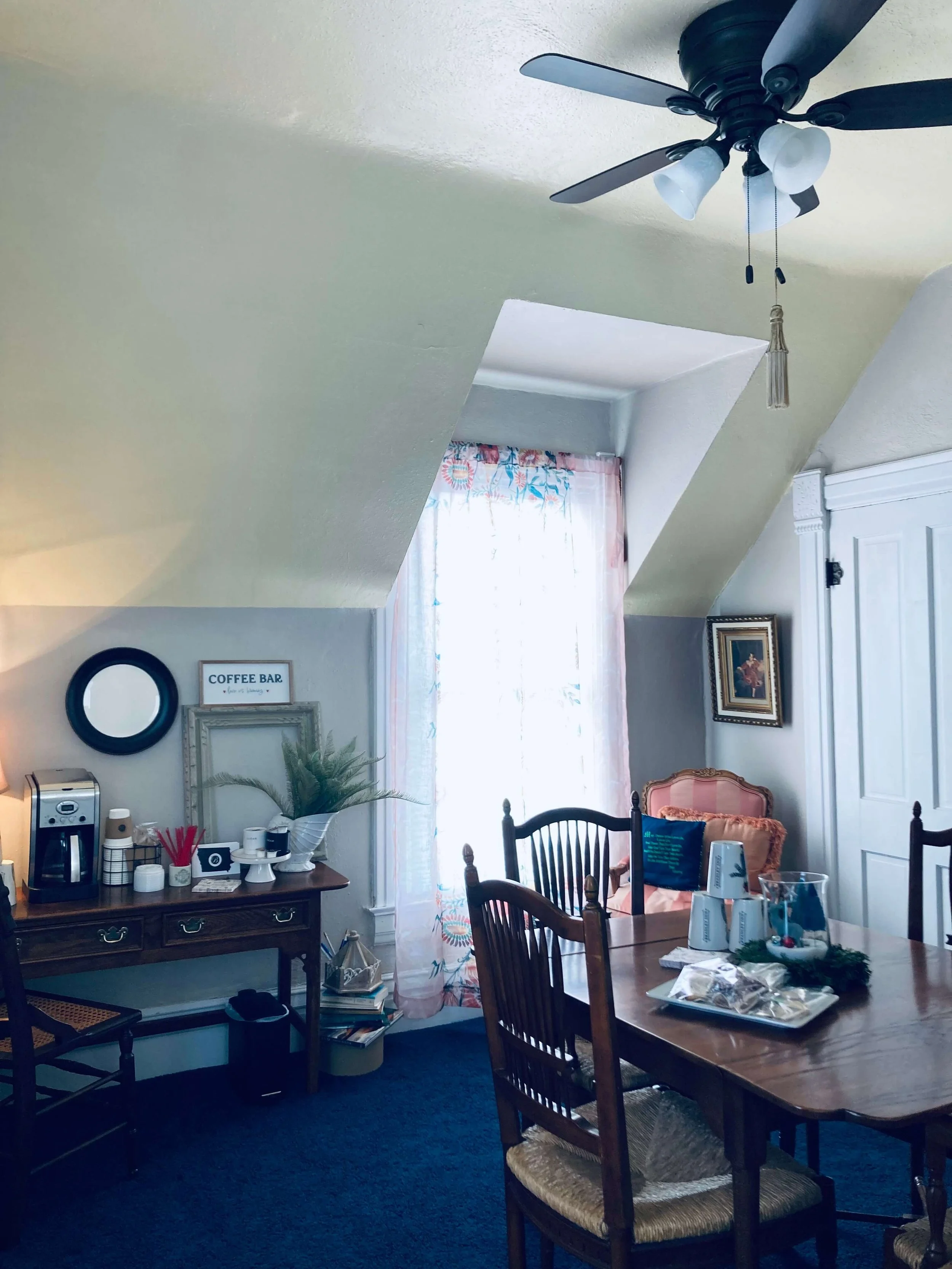 A cozy dining room with a wooden table, chairs, a ceiling fan, a window with floral curtains, and a sideboard with coffee-making supplies and decorative items.
