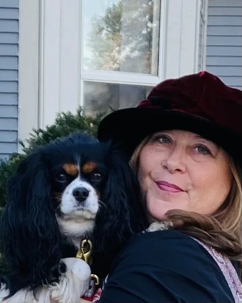 A woman with light-colored hair wearing a dark red hat, smiling and posing for a photo beside a black and white dog with long floppy ears. They are outdoors in front of a house with a window and some greenery.