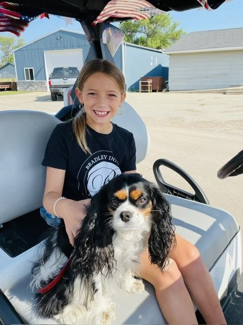A girl with a big smile sitting in a golf cart with a black and white dog on her lap outside on a sunny day.
