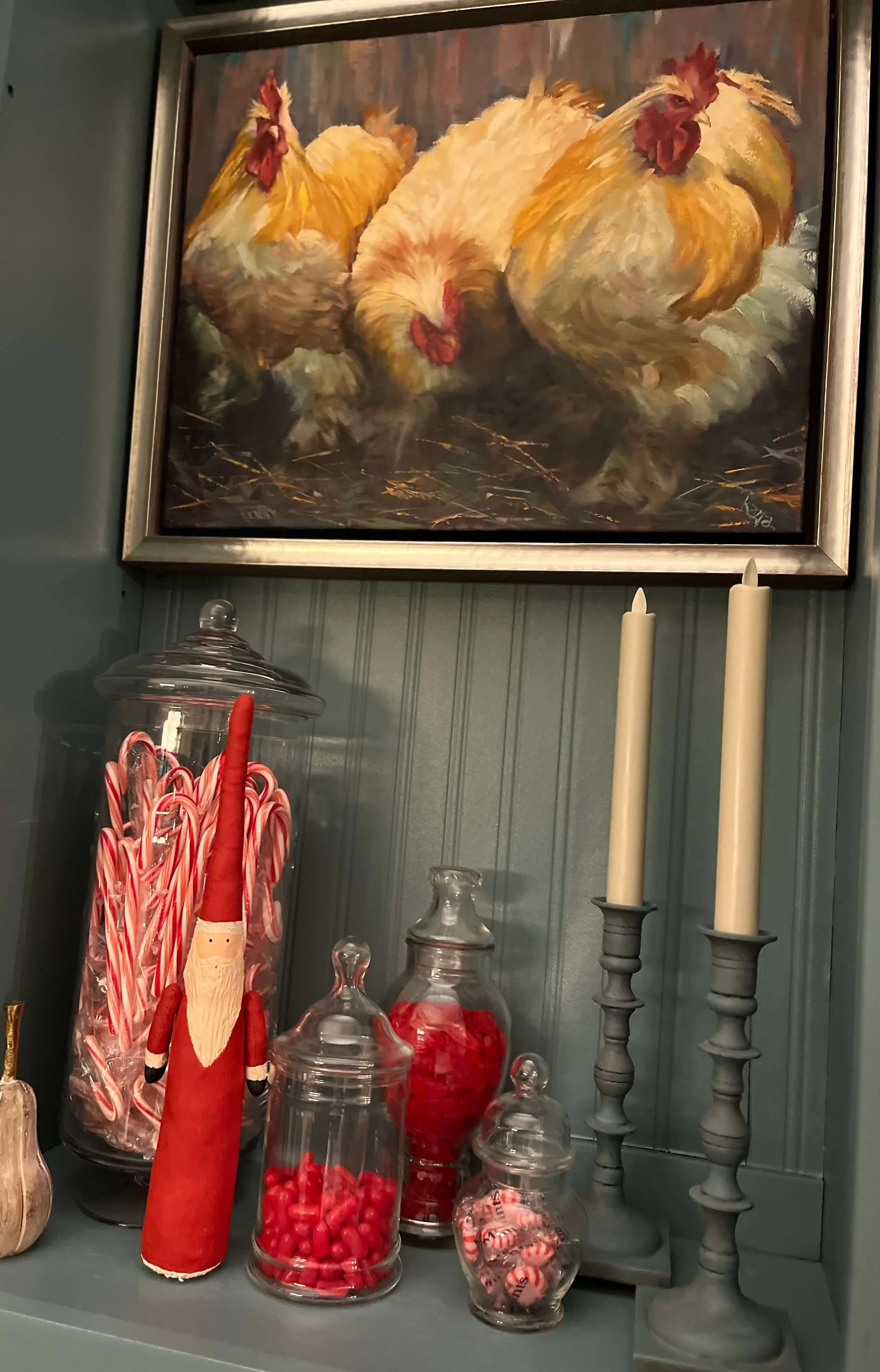 A decorative shelf with a painting of three chickens hanging above it. The shelf holds various glass jars filled with red candies and a gnome doll dressed as Santa Claus, along with two tall gray candlesticks with white candles.