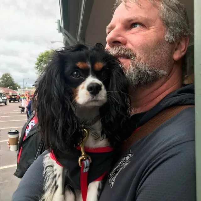 A middle-aged man with gray hair and beard holding a black, brown, and white Cavalier King Charles Spaniel dog outside a building.