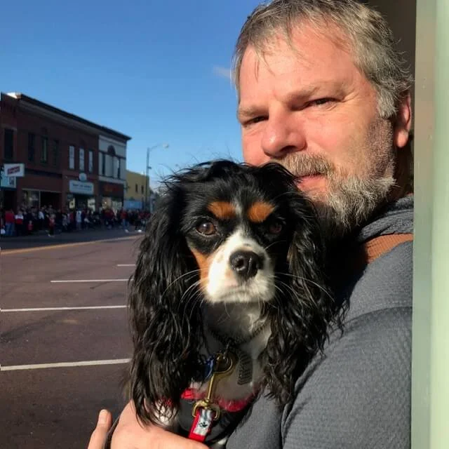 A man with gray hair and beard holding a black, brown, and white Cavalier King Charles Spaniel in front of a storefront on a busy street.