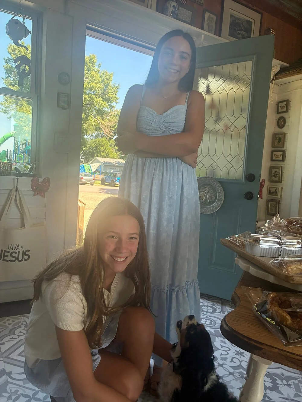 Two young girls, one standing in a doorway and smiling, the other sitting on the floor and smiling, with a puppy, in a cozy home with decorated walls and a table with snacks.