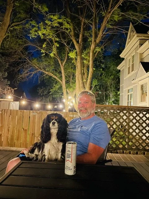 A man with gray hair and beard smiling while sitting at a table outdoors at night, with a black and white dog sitting on the table. There are string lights hanging in the background, a wooden fence, a large tree, and a white house with multiple windo