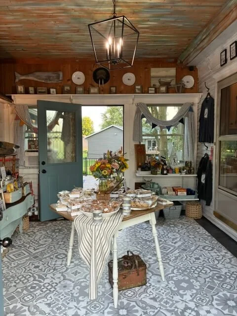 A cozy kitchen with a rustic wooden ceiling, patterned tile floor, and a white table set for a meal with a floral centerpiece. The room has a large door and window with curtains, open to a backyard with trees. Shelves and wall decor include vintage a