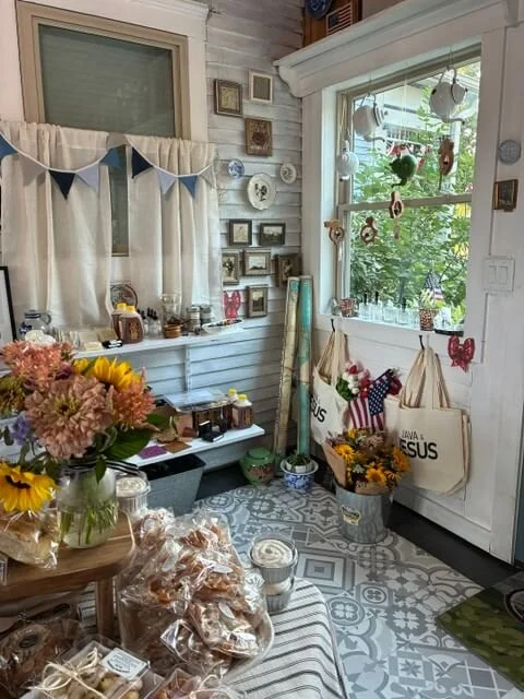 A cozy sunlit corner of a room decorated with flowers, hanging bags, and various decorative items on white paneled walls and window.
