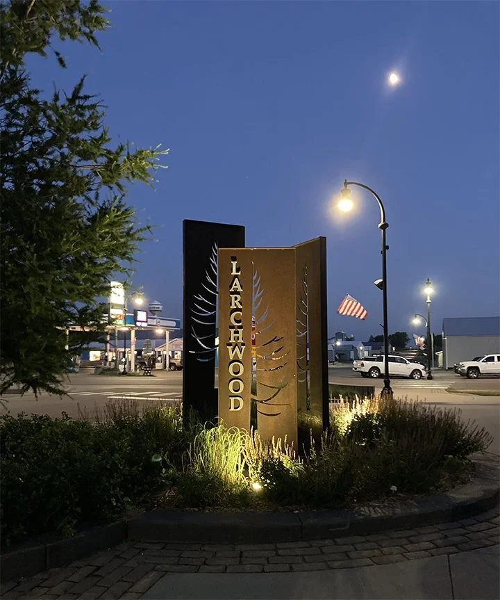 Street scene in the evening showing a sign that reads 'LARCHWOOD' illuminated by ground lights. In the background, a gas station, parked cars, and a partially visible American flag are visible. Street lamps are lit, and the moon is visible in the cle