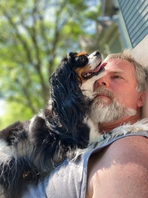 A man lying outside with a Cavalier King Charles Spaniel dog resting its head on his face, outdoor greenery in background.