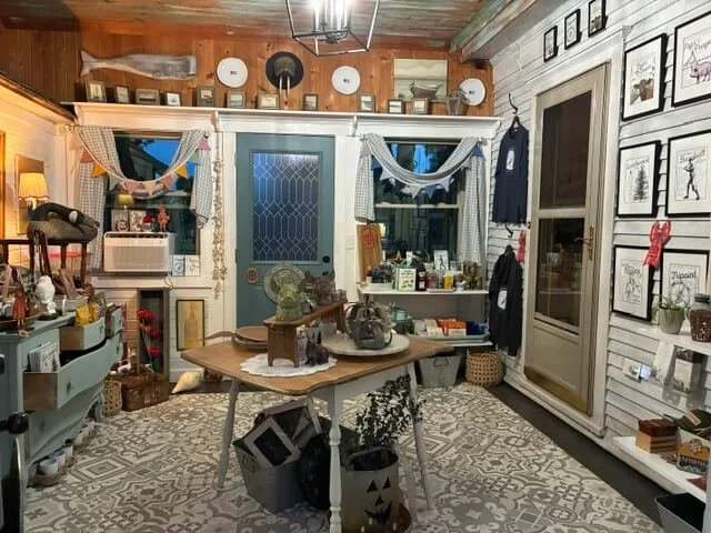 Cozy vintage-style room with wooden walls and ceiling, decorated with framed artwork, curtains, and various knick-knacks. Contains a table with decorative items, baskets, and a patterned rug.