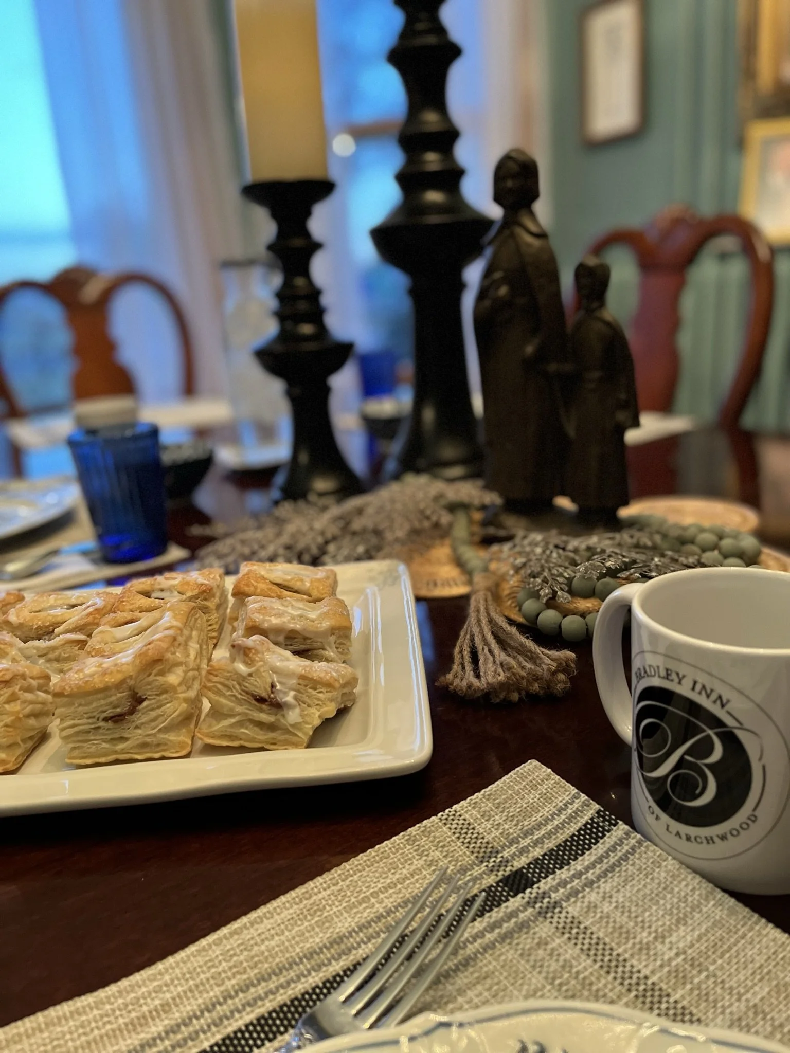 A table set with a plate of layered pastry squares, a white mug with a black logo, multiple black candles, decorative statues of two figures, and jewelry, with a dining room with a window and curtains in the background.