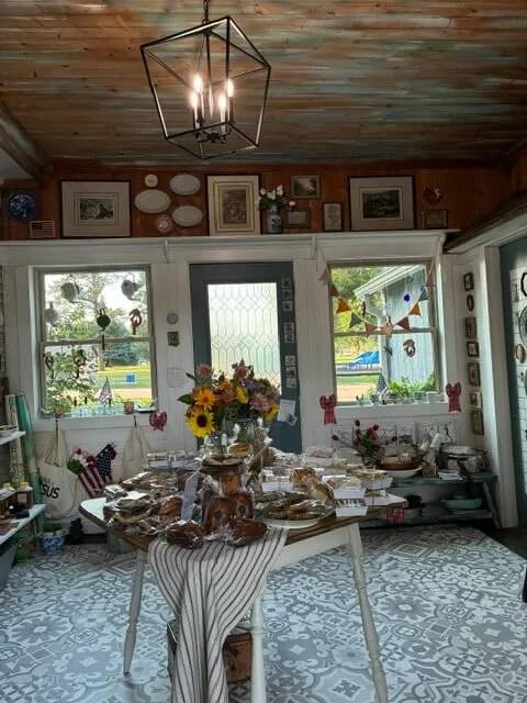 Indoor dining area with a round table covered with a white and brown striped tablecloth, set with food and decorations, sunflower centerpiece, large windows, wooden ceiling, wall art, and a modern black geometric chandelier.