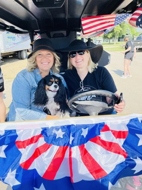 Two smiling women sitting in a decorated golf cart with a dog, holding American flags, celebrating the Fourth of July.