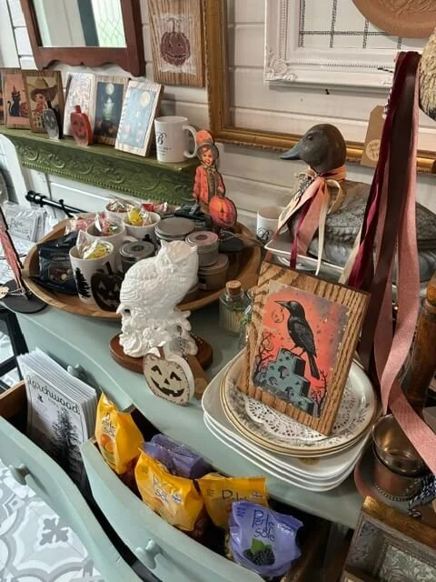A tabletop decorated with various Halloween-themed items including a white ceramic skull, a pumpkin, a painted crow on a wooden plaque, and small figurines. There are also packaged snacks, candles, framed photographs, and decorative objects in the ba