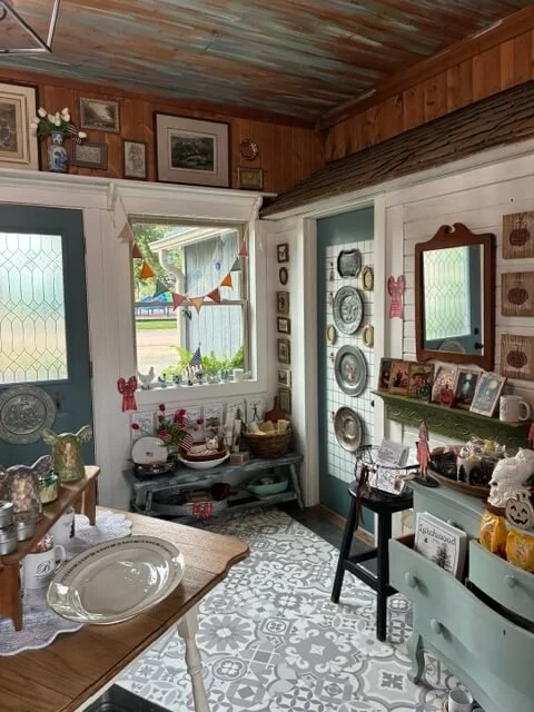 Cozy vintage-style room with wooden walls, decorated with pictures, plates, and ornaments, featuring a window with bunting, a small table with decorative items, and a mirror on the wall.