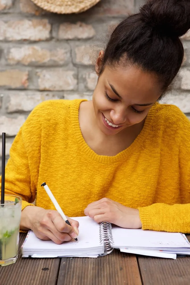 Image shows a young woman in a bright yellow sweater writing in a notebook and sitting at a table outside.