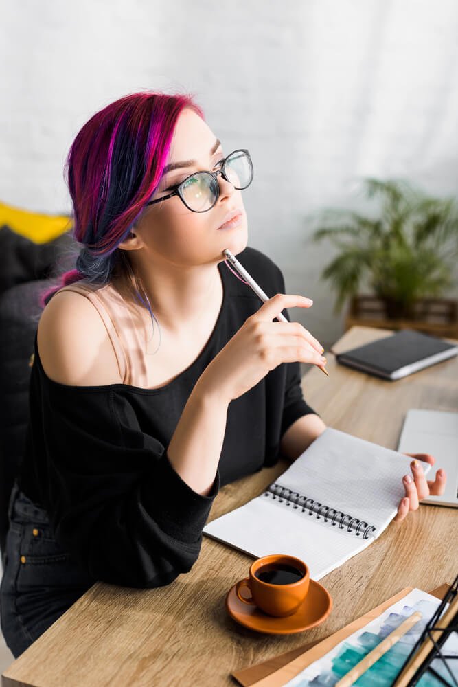 Image of a woman with purple hair sitting with a notebook and staring off into the distance.