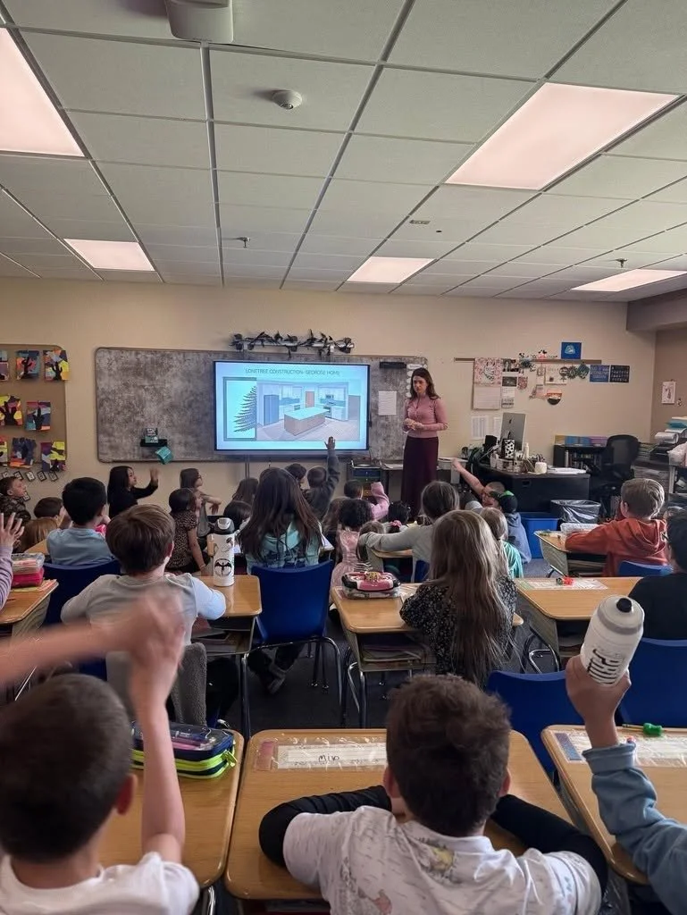 A classroom filled with young students sitting at desks, raising their hands, while a teacher stands near a whiteboard with a projected image of a classroom interior.