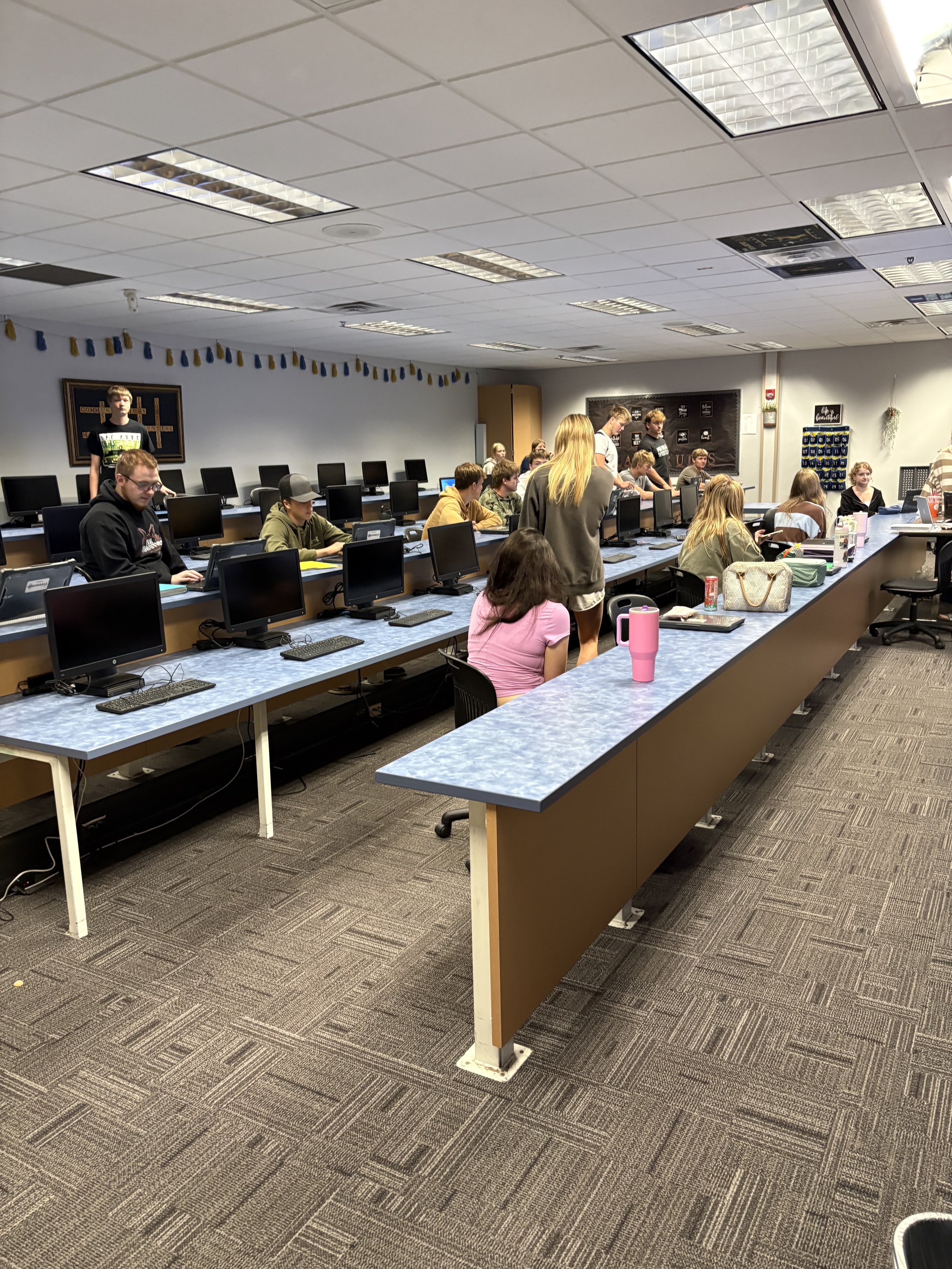 A classroom or computer lab with multiple desks arranged in a U-shape, each with a computer monitor and keyboard. Several students are seated and working on computers, while a few others are standing and assisting. The room has a white ceiling with fluorescent lights, and decorations including banners and framed posters on the walls.