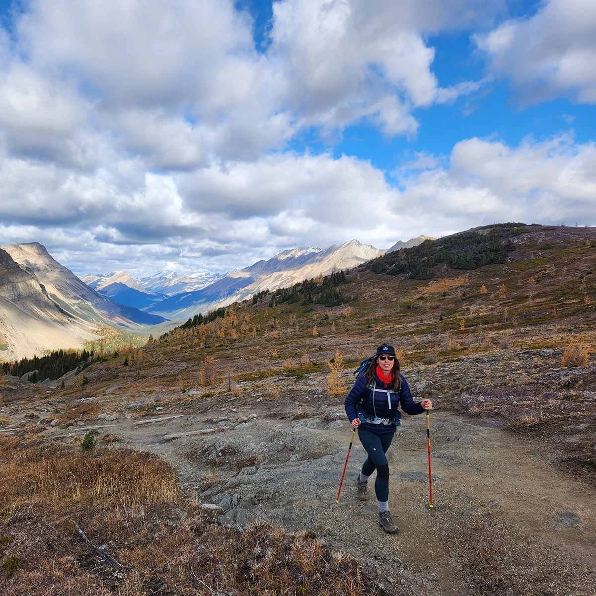 A woman hiking on a trail in a mountainous landscape with snow-capped peaks, cloudy sky, and autumn foliage in the background