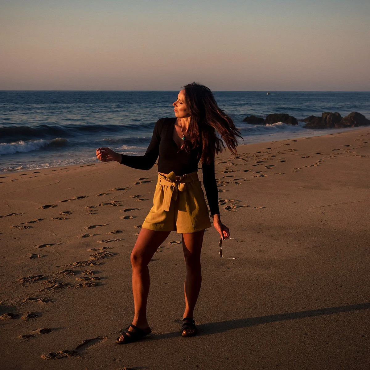 Woman standing on a sandy beach at sunset, facing sideways with hair blowing in the wind, holding sunglasses in one hand, with ocean waves and rocks in the background.