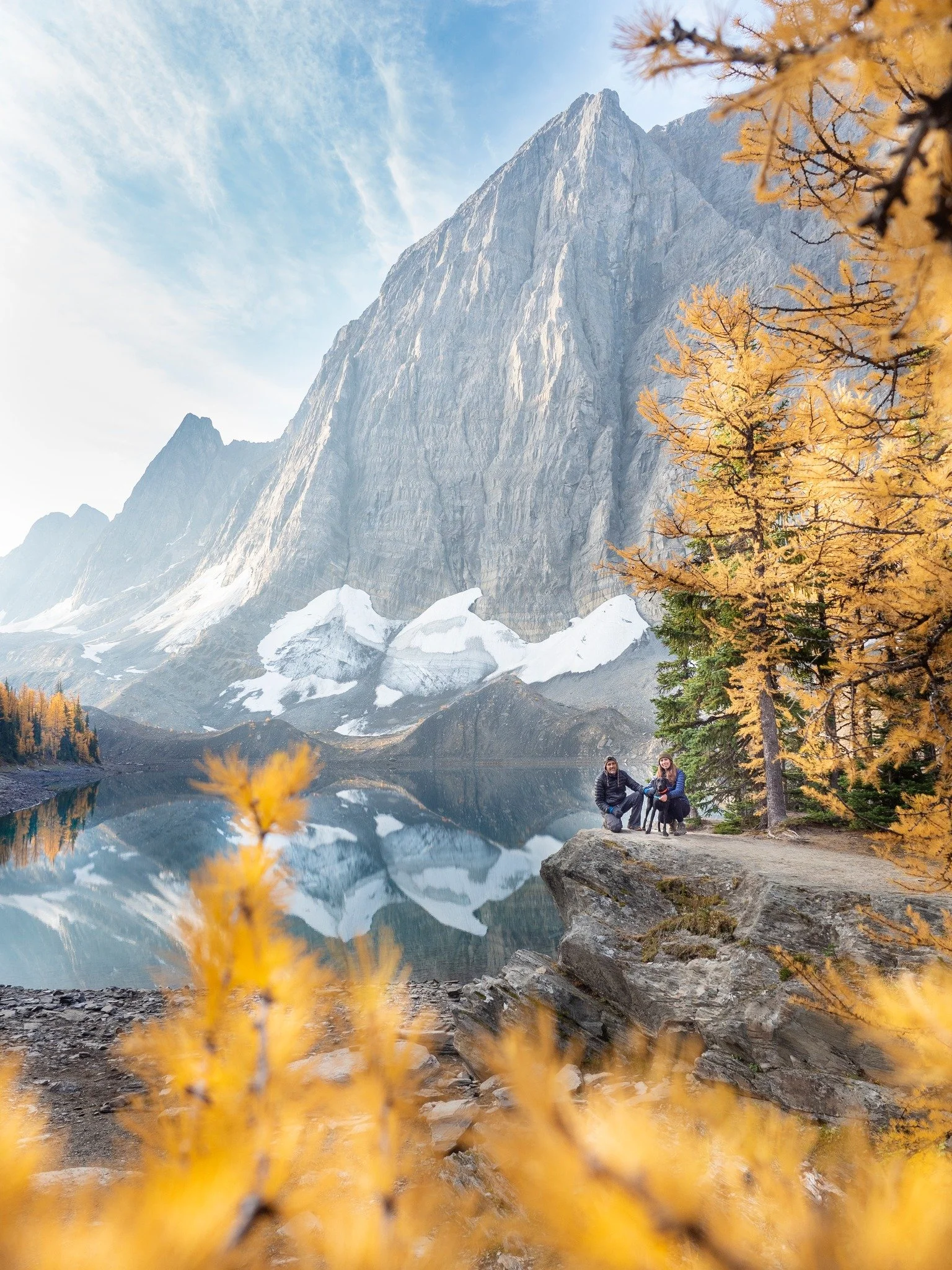 Two people sitting on a rock by a lake with a mountain and snow in the background, surrounded by yellow-orange fall trees in the Rocky Mountains or Alberta