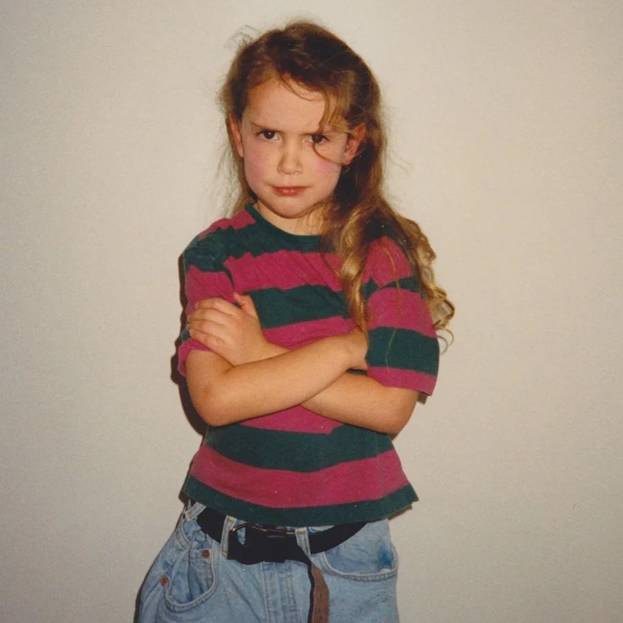 A young girl with wavy light brown hair and a serious expression, crossing her arms, standing against a plain wall.